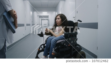 Hospital corridor: Woman with SMA in wheelchair talks to doctor 107012462