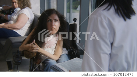 Woman with SMA in wheelchair talks to doctor in hospital lobby 107012475