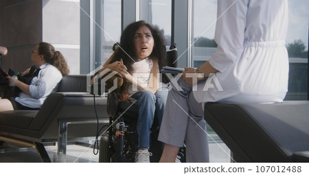 Woman with SMA in wheelchair talks to doctor in hospital lobby 107012488