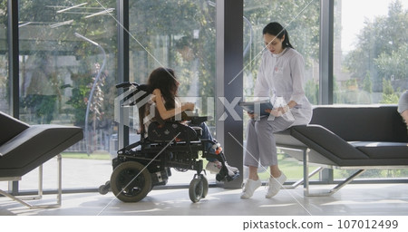 Female doctor consults patient with physical disability in clinic lobby 107012499