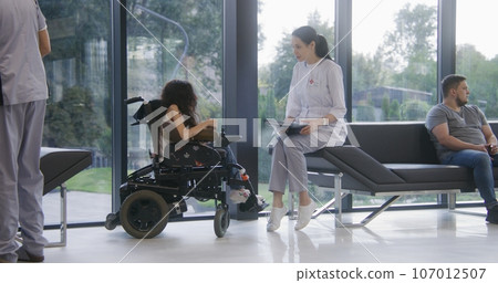 Female doctor consults patient with physical disability in clinic lobby 107012507