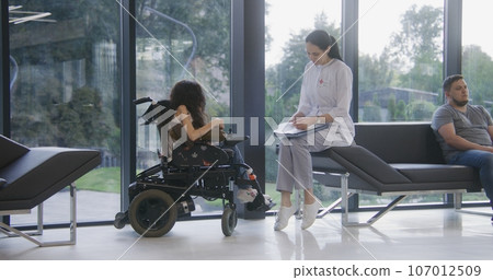 Female doctor consults patient with physical disability in clinic lobby Female doctor consults patient with physical disability in clinic lobby 107012509