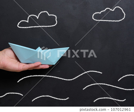 A female hand holds a paper boat against a backdrop of a board with drawn waves A female hand holds a paper boat against a backdrop of a board with drawn waves 107012961