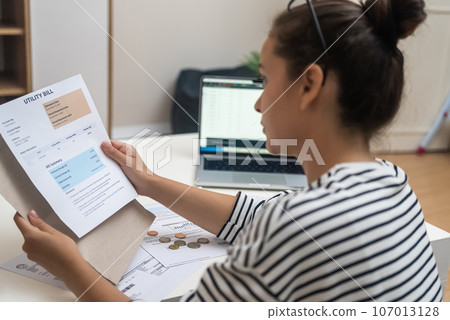 Woman with hair bun looks at utility bills with total amount for online payment through laptop at workplace in office housewife examines data in bank documents at home 107013128