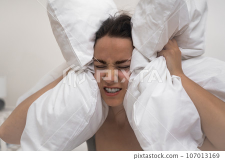 Young woman suffers from insomnia sick female presses white pillows to head frowning and clenching teeth person with headache and sleep disorders closeup 107013169
