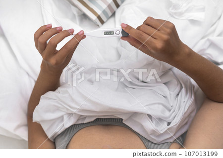 An unwell woman, in bed, assesses her body temperature with a thermometer, tissues on hand, facing the challenges of fever and sickness.  107013199