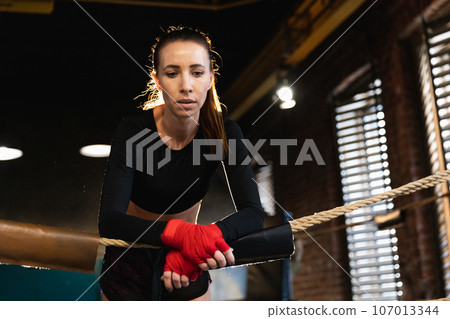 Woman fighter girl power. Woman fighter with red boxing wraps protective bandages standing on boxing ring leaning on ropes waiting resting. Strong powerful girl. Strength fit body workout training 107013344