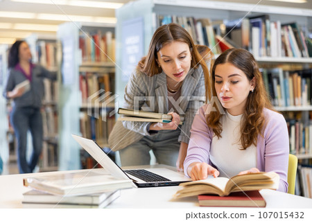 Two female students studying in library with books and laptop Two female students studying in library with books and laptop 107015432