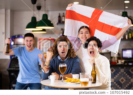 Enthusiastic company of young adult sports fans waving flag of England and supporting national team with beer in pub Enthusiastic company of young adult sports fans waving flag of England and supporting national team with beer in pub 107015492