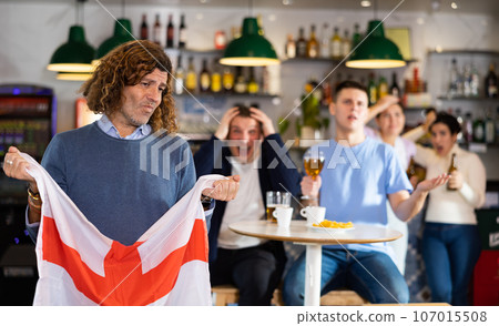 Company of young adult sports supporters with national flag of England upset about favorite team losing match in bar Company of young adult sports supporters with national flag of England upset about favorite team losing match in bar 107015508