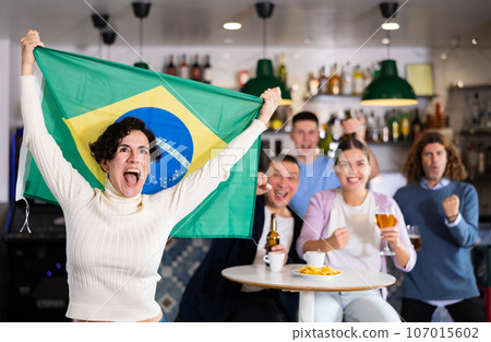 Company of emotional young adult fans supporting sports team of Brazil with state flag while resting with beer in bar Company of emotional young adult fans supporting sports team of Brazil with state flag while resting with beer in bar 107015602