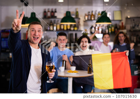Group of friends sports supporters cheering for favorite team of Belgium with flag while watching match on TV in pub Group of friends sports supporters cheering for favorite team of Belgium with flag while watching match on TV in pub 107015630