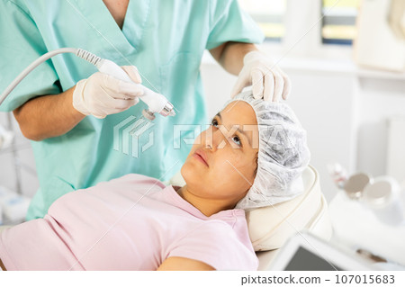 Young male cosmetologist doing moisturizing procedure to young woman 107015683