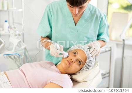 Young male cosmetologist doing ultrasound procedure to young woman Young male cosmetologist doing ultrasound procedure to young woman 107015704