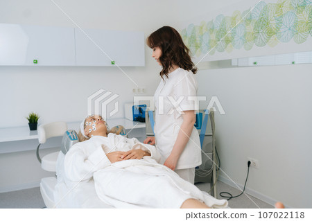 Remote view of friendly female physiotherapist communicating with lying young woman client during facial myostimulation procedure in modern beauty clinic. Concept of non-surgery cosmetology. Remote view of friendly female physiotherapist communicating with lying young woman client during facial myostimulation procedure in modern beauty clinic. Concept of non-surgery cosmetology. 107022118