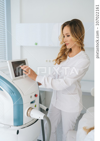 Vertical shot of professional female cosmetologist adjusts device for photorejuvenation using touchscreen in beauty clinic. Concept of non-surgery cosmetology, professional skincare, beauty treatment. 107022161