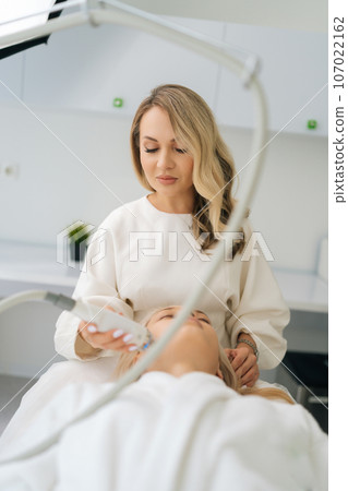 Vertical portrait of focused cosmetologist making woman RF lifting procedure of face and eyes area. Female client getting stimulating beauty facial treatment lying on couch in beauty clinic. 107022162