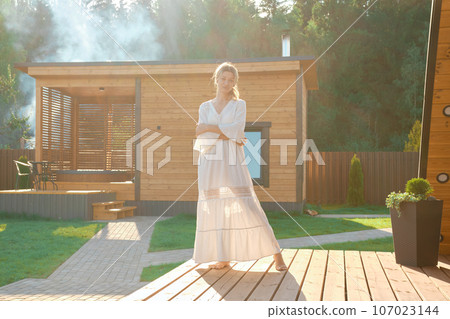 Dreamy woman in long white dress standing on terrace with hot tub and sauna on background 107023144