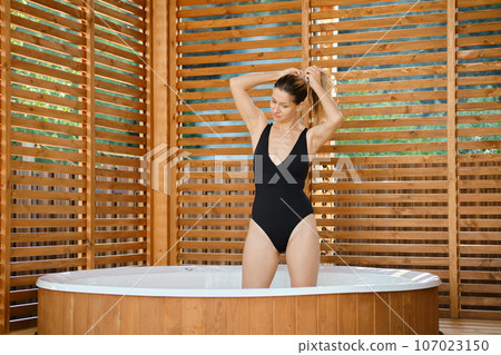 Woman standing in hot tub in a swimsuit and collects her hair in a ponytail Woman standing in hot tub in a swimsuit and collects her hair in a ponytail 107023150