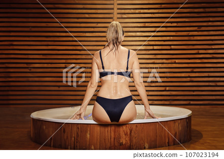 Rear view of a woman in a bikini sitting on the edge of a hot tub in the evening 107023194