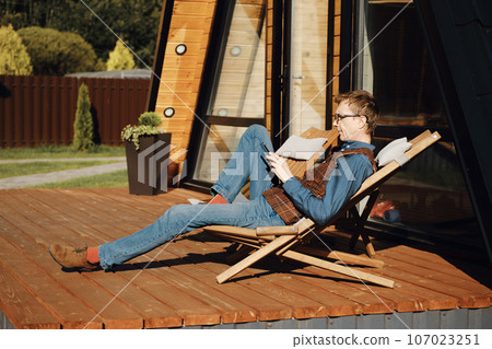 Middle aged man in brown vest reading message on smartphone sitting on terrace of tiny house 107023251