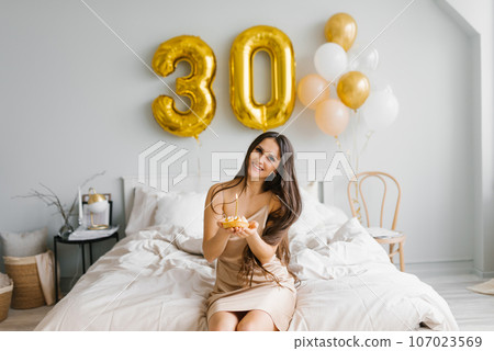 Happy woman with long hair is sitting on a bed in a cozy bedroom at home with a cake on a plate and a candle and celebrating her thirtieth birthday 107023569