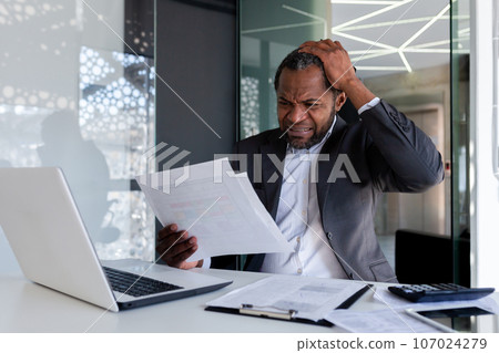 Frustrated and angry financier at workplace inside office, angry boss reviewing invoices contracts and reports sitting at table with laptop, businessman behind paper work. 107024279