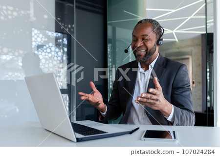 Mature experienced man with headset phone inside office at workplace, smiling talking remotely and advising customers, african american customer support worker, using video call and laptop. 107024284