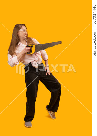 Young female model with hand saw posing for photo in studio against yellow background. Strong modern woman with man tools. Vertical frame. Copy space. 107024440