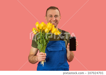 Portrait of smiling flower delivery man holding smartphone with blank black screen on pink background. Order flowers online in mobile application. 107024701