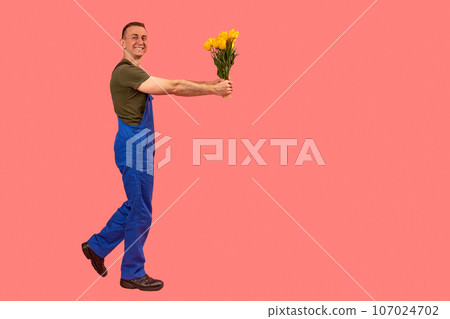 Caucasian male courier holding bouquet. Flower delivery concept. Man in workers overalls holds out flowers. Side view. Copy space, mock up. Caucasian male courier holding bouquet. Flower delivery concept. Man in workers overalls holds out flowers. Side view. Copy space, mock up. 107024702