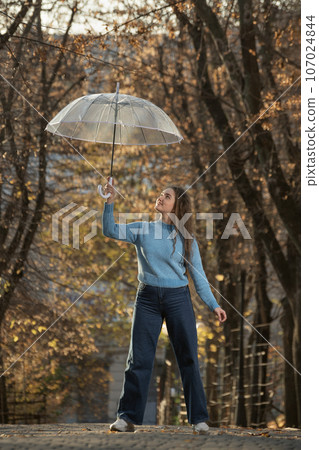 Happy young woman with long hair walks in autumn park. Beautiful girl with transparent umbrella Happy young woman with long hair walks in autumn park. Beautiful girl with transparent umbrella 107024844