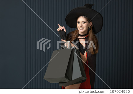 Happy smiling woman in witch hat after shopping with black shopping bags pointing up. Dark blue background. Halloween sale. Seasonal discounts Happy smiling woman in witch hat after shopping with black shopping bags pointing up. Dark blue background. Halloween sale. Seasonal discounts 107024930
