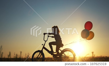girl kid silhouette bike riding on a park. kid girl rides a bike in nature in the park on the road. happy family kid dream concept. daughter plays a bike lifestyle rides on a sandy road girl kid silhouette bike riding on a park. kid girl rides a bike in nature in the park on the road. happy family kid dream concept. daughter plays a bike lifestyle rides on a sandy road 107025319