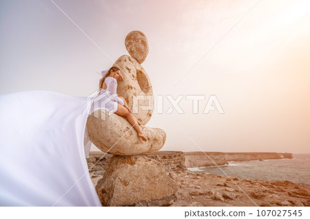 Woman sky stone. Lady stands on stone sculpture with ocean view. She is dressed in a white long dress, against the backdrop of the sea and sky. The dress develops in the wind. Woman sky stone. Lady stands on stone sculpture with ocean view. She is dressed in a white long dress, against the backdrop of the sea and sky. The dress develops in the wind. 107027545