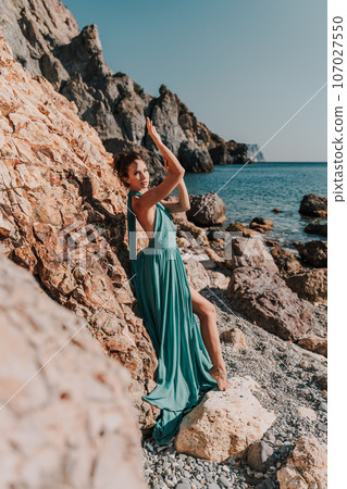 Woman green dress sea. Woman in a long mint dress posing on a beach with rocks on sunny day. Girl on the nature on blue sky background. Woman green dress sea. Woman in a long mint dress posing on a beach with rocks on sunny day. Girl on the nature on blue sky background. 107027550