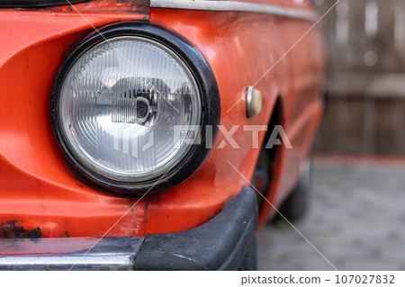 Red old Zaporozhets car in the yard on a sunny day. The car has round headlights. Red old Zaporozhets car in the yard on a sunny day. The car has round headlights. 107027832