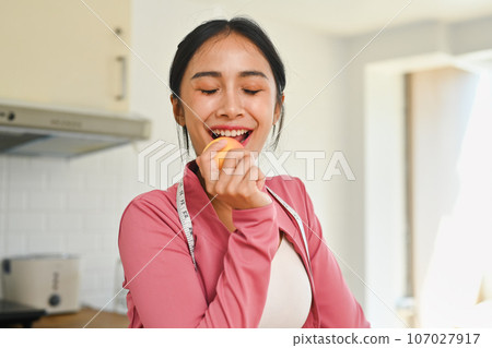 Charming young woman in sportswear eating an apple after exercising at home. Healthy food concept Charming young woman in sportswear eating an apple after exercising at home. Healthy food concept 107027917