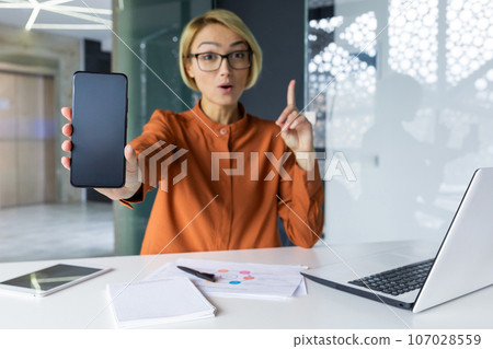 Joyful and successful business woman shows phone screen to camera, blank space for recording. Woman at workplace recommending financial app using smartphone inside office. Joyful and successful business woman shows phone screen to camera, blank space for recording. Woman at workplace recommending financial app using smartphone inside office. 107028559