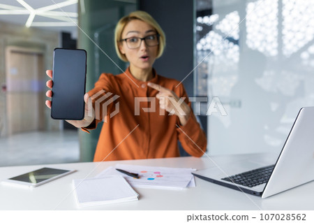 Joyful and successful business woman shows phone screen to camera, blank space for recording. Woman at workplace recommending financial app using smartphone inside office. 107028562