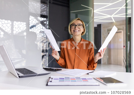 Joyful and successful businesswoman financier happy with achievement result, celebrating victory at workplace inside office, female worker holding contract reports and bills in hands. Joyful and successful businesswoman financier happy with achievement result, celebrating victory at workplace inside office, female worker holding contract reports and bills in hands. 107028569