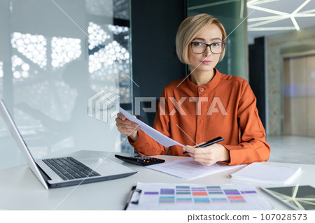 Serious and pensive business woman behind paper work inside office, female financier worker thinks about contracts and reports with charts and graphs, blonde successful woman uses laptop at work. Serious and pensive business woman behind paper work inside office, female financier worker thinks about contracts and reports with charts and graphs, blonde successful woman uses laptop at work. 107028573