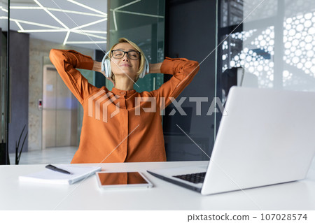Young beautiful businesswoman relaxing in the office listening to music in headphones, female programmer with her hands behind her head smiling with closed eyes, well done worker completing project. 107028574
