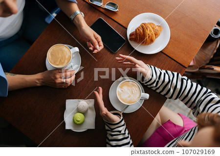 Closeup high-angle view of two unrecognizable happy female girlfriends sitting at table with coffee, croissant and macaroon in cafe, communicate and sharing stories spending relax in coffee shop. Closeup high-angle view of two unrecognizable happy female girlfriends sitting at table with coffee, croissant and macaroon in cafe, communicate and sharing stories spending relax in coffee shop. 107029169