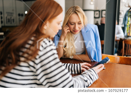 Rear view of two pretty young women sitting at table with coffee at cafe by window, having conversation and showing each other something on mobile phones. Concept of woman friendship 107029177