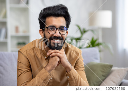 Close-up portrait of young smiling hispanic man smiling and looking at camera while sitting on sofa in living room. Close-up portrait of young smiling hispanic man smiling and looking at camera while sitting on sofa in living room. 107029568