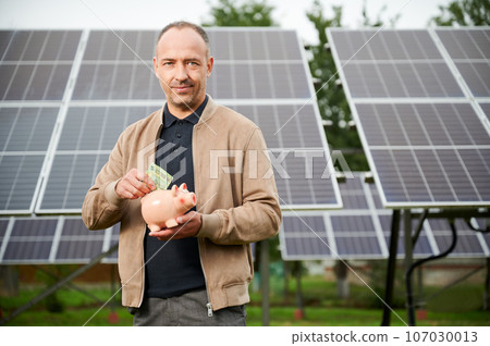 Confidence man putting earned money to piggy bank. Male adult saving profit he got from investing in green energy. Man in casual clothes holding moneybox and putting euros in it. Confidence man putting earned money to piggy bank. Male adult saving profit he got from investing in green energy. Man in casual clothes holding moneybox and putting euros in it. 107030013