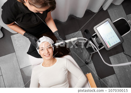 Top view of smiling woman lying near cosmetology equipment and receiving facial treatment. Cosmetologist using radiofrequency microneedling device while performing beauty skincare procedure. Top view of smiling woman lying near cosmetology equipment and receiving facial treatment. Cosmetologist using radiofrequency microneedling device while performing beauty skincare procedure. 107030015