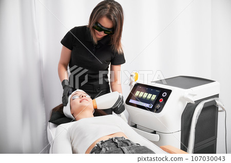 Cosmetologist in safety glasses using diode laser device while performing facial skincare procedure. Woman lying near cosmetology equipment and receiving face rejuvenation treatment in beauty salon. Cosmetologist in safety glasses using diode laser device while performing facial skincare procedure. Woman lying near cosmetology equipment and receiving face rejuvenation treatment in beauty salon. 107030043