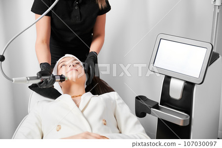 Young woman lying near cosmetology equipment and receiving facial treatment in beauty salon. Female cosmetologist performing skincare procedure with radiofrequency microneedling device. 107030097